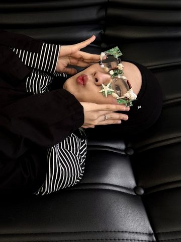 Anika lying on a black leather sofa, wearing glasses decorated with shells and circuit boards